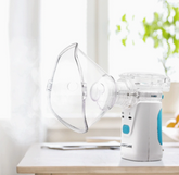 An Alertcare Portable Ultrasonic Nebulizer HL100R by Faire.com rests on a table near a window with daylight streaming in and a green plant in the background.