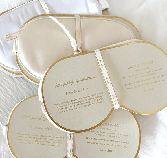 A set of Satin Sleep Masks by LoveLina, adorned in a white and gold color scheme, on a cozy white bed. (Available on Faire.com)