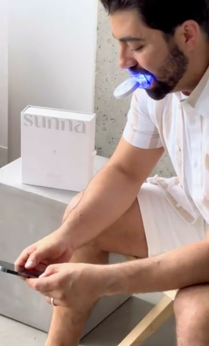 A man diligently performing his home whitening routine, sitting in front of a box containing an Advanced Home Whitening Kit by Sunna toothbrush.