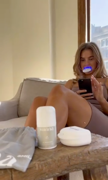 A woman sitting on a couch looking at her phone while using the Advanced Home Whitening Kit by Sunna for her whitening routine.